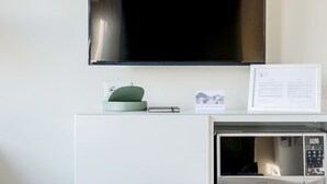 A hotel room with a flat-screen TV mounted on the wall, a microwave oven, a small table with a green object and a notebook, and a 'No Smoking' sign. - Mono A (Milan)