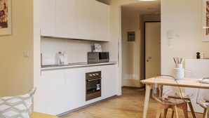 A compact kitchen with white cabinets, a microwave, and an oven. There's a wooden table with chairs and a small white cabinet with utensils. - Mono B (Milan)