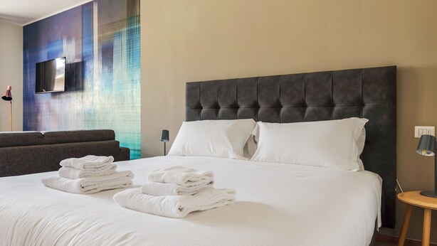 A hotel room with a large bed, a flat-screen TV mounted on the wall, a grey sofa, and a small table with a lamp. - Mono B (Milan)