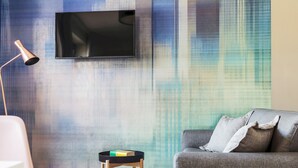 A modern living room with a mounted TV, a floor lamp, a small side table with books, and a grey sofa with cushions. - Mono B (Milan)
