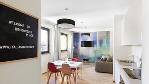 A modern living room with a dining area, a sofa, and a kitchen. There is a blackboard with "WELCOME TO BERGONZOLI PLACE" and a website address. - Mono B (Milan)