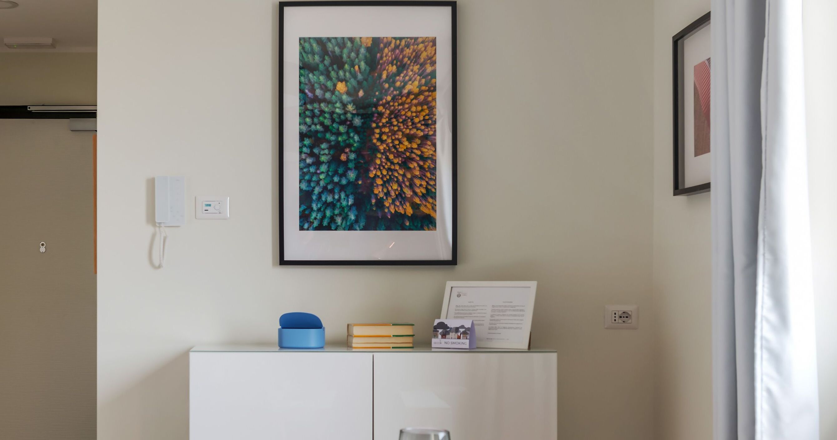 A framed artwork on a wall, a white cabinet with a blue object and books, and a framed certificate on top.
