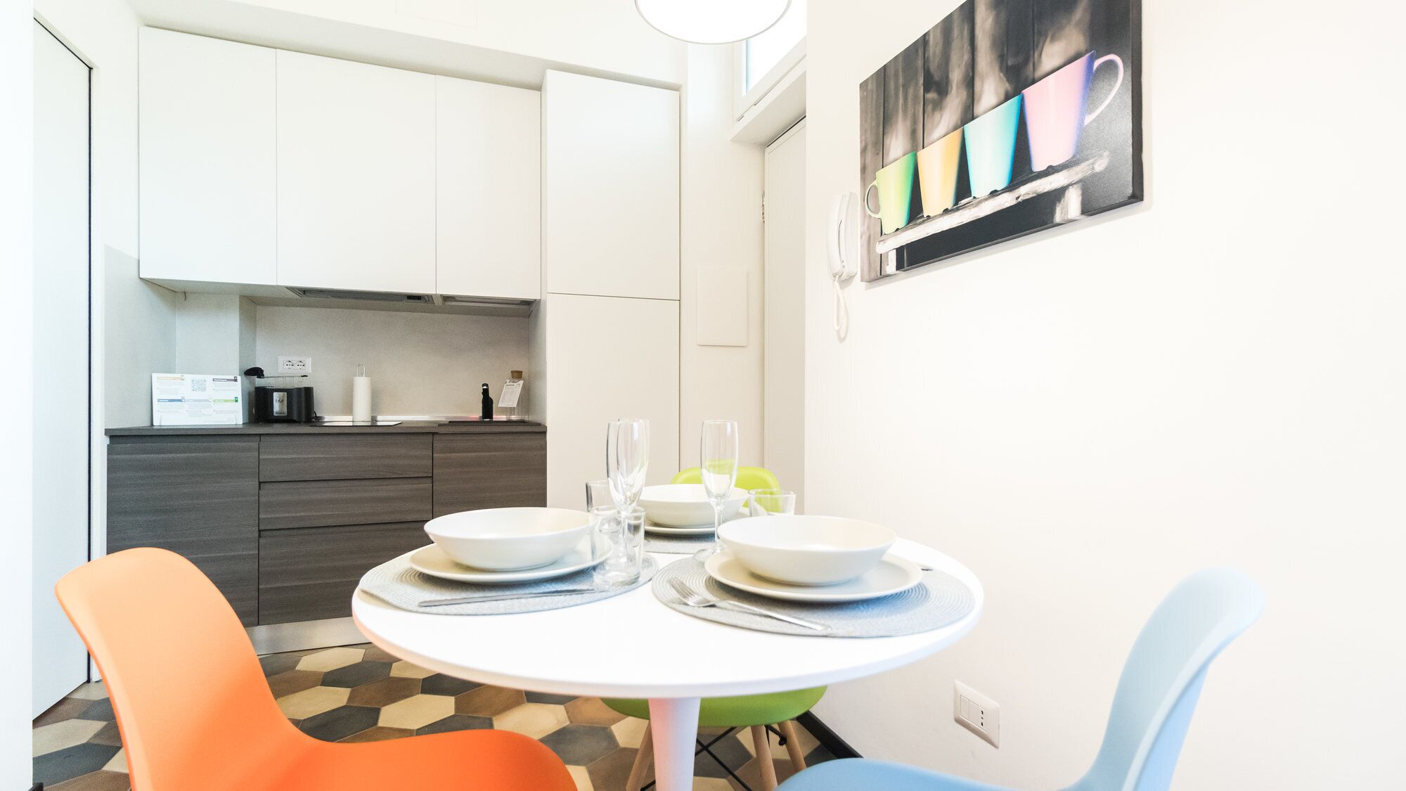 A compact kitchen with white cabinets, a dining table set with plates and glasses, and a colorful painting on the wall.