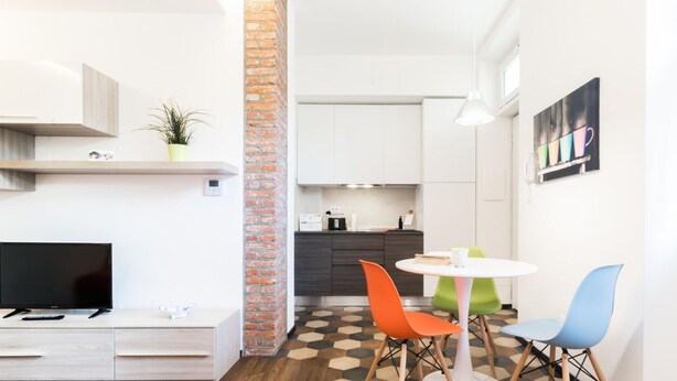 A modern apartment with a TV, a dining area with colorful chairs, a kitchen, and a brick accent wall. - Italianway Easy - Ciaia 6 B (Milan)