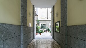 A corridor with tiled flooring, flanked by walls with a stone pattern, leading to a courtyard with potted plants and a bicycle. - Mi-clod9a3 - Corso Lodi 9 (Milan)