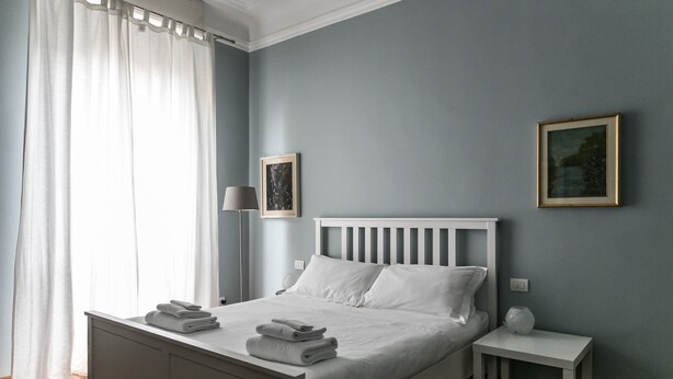 A neatly made bed with white linens and folded towels, a bedside table with a lamp, and a framed picture on the wall. - Italianway Easy - Da Procida 25 (Milan)