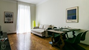 A modern living room with a sofa, a dining table set with glasses, a lamp, and a framed picture on the wall. - Italianway Easy - Da Procida 25 (Milan)