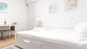 Ein minimalistisches Schlafzimmer mit Bett, Nachttisch, Lampe und Schreibtisch mit Stühlen.