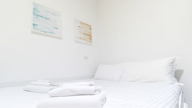 Une chambre d’hôtel propre et blanche, avec un lit bien fait, deux œuvres d’art accrochées au mur et une petite table de chevet avec une lampe.