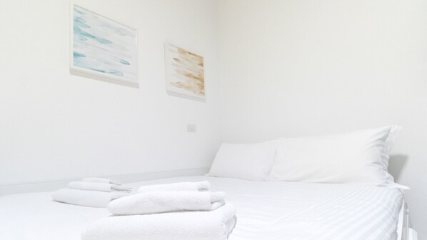 A clean, white hotel room with a neatly made bed, two wall-mounted artworks, and a small bedside table with a lamp.