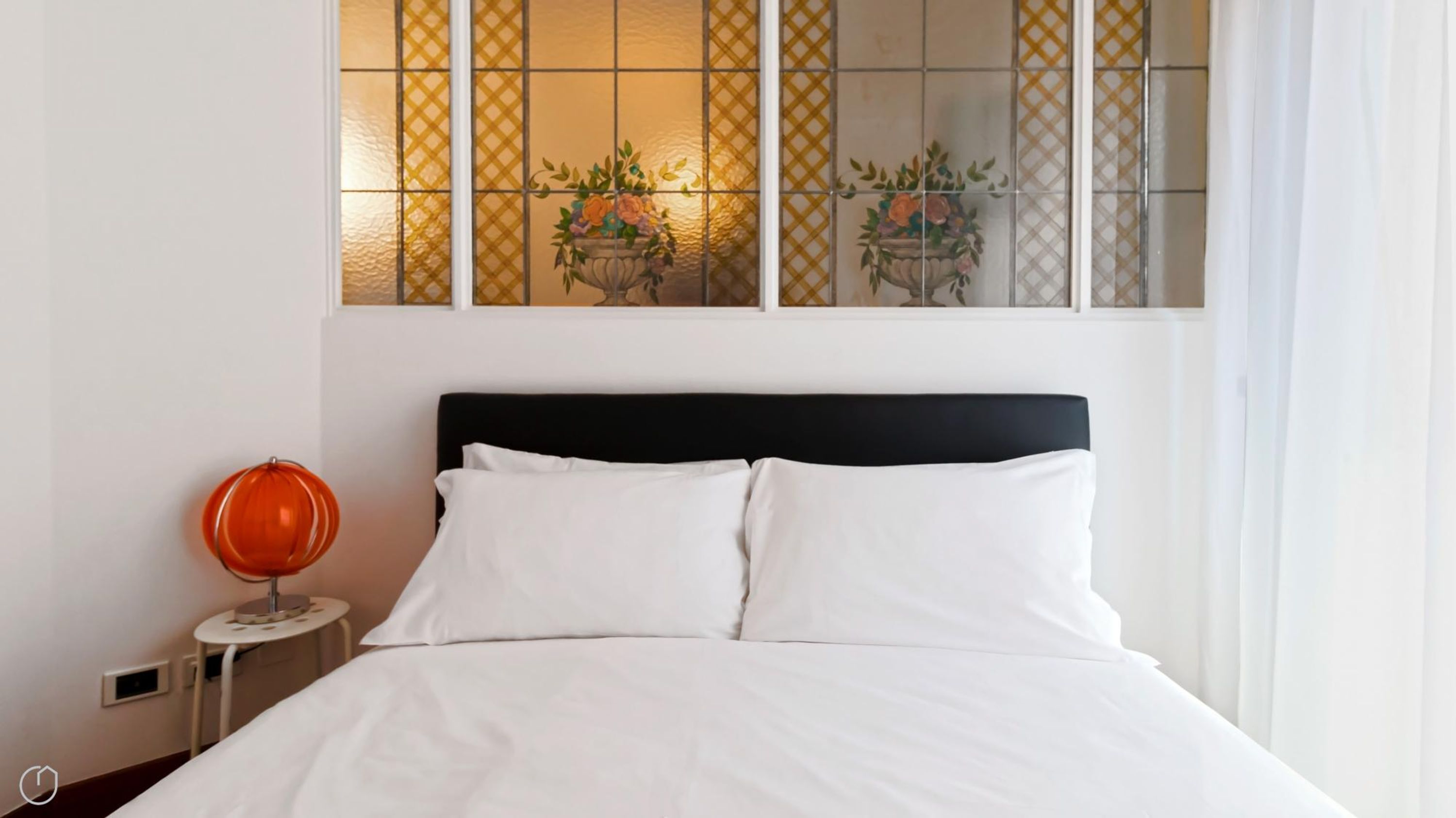 Ein Bett mit weißen Leinentüchern, ein schwarzer Kopfteil und ein kleiner runder Tisch mit einer roten Lampe daneben.