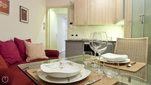 A modern living room with a glass-top dining table set for two, featuring white plates with floral designs. A wicker chair and a red sofa with cushions are visible. The kitchen in the background has wooden cabinets and a small island. - Italianway Easy - Fiori Chiari 20 (Milan)