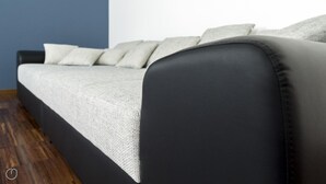 Un sofá moderno con tapicería de color gris claro y reposabrazos de cuero negro, situado frente a una pared azul y suelo de madera.