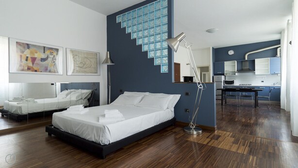 Una habitación de hotel moderna con dos camas, un ventanal con bloques de vidrio azul, una kitchenette y piso de madera.