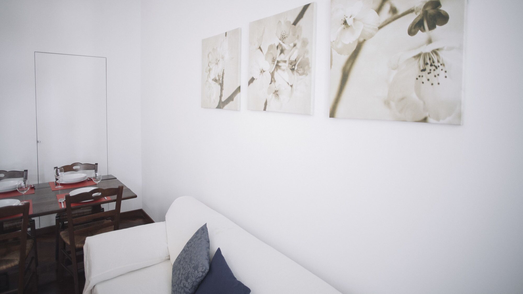 Un comedor con una mesa blanca dispuesta para cuatro personas, sillas de madera y un sofá blanco con un cojín azul.