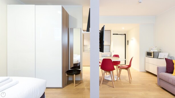 Ein modernes Hotelzimmer mit Bett, Schrank, einer kleinen Küchenzeile mit Mikrowelle und einem Esstisch mit roten Stühlen.
