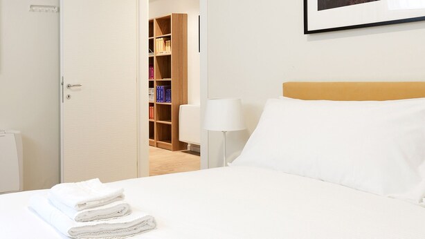 A hotel room with a neatly made bed, a bedside lamp, and a wooden bookshelf with books.