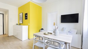 Een heldergele muur, een wit bureau met stoelen, een witte bank en een flatscreen televisie.