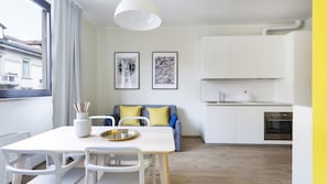 Une cuisine moderne avec des armoires blanches, un évier en acier inoxydable et un four. On y trouve une table à manger avec des chaises blanches et un petit vase blanc contenant des crayons. Deux tableaux encadrés sont accrochés au mur, au-dessus du canapé.