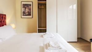 A hotel room with a bed, a red headboard, a framed picture on the wall, a closet with hanging clothes, and a folded towel on the bed. - Mi-brgz1c2 Bergonzoli Place - Bilo B - Blues (Milan)
