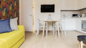 A compact living space with a yellow sofa, a dining table with chairs, a TV mounted on the wall, a microwave, and a stove in the kitchen area. - Mi-brgz1c2 Bergonzoli Place - Bilo B - Blues (Milan)