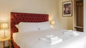 A hotel room with a tufted red headboard, white bedding, two bedside lamps, and a neatly folded stack of towels on the bed.