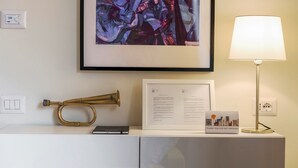 A hotel room with a framed abstract painting, a brass trumpet, a framed document, a thank you card, a lamp, and a power outlet. - Mi-brgz1c2 Bergonzoli Place - Bilo B - Blues (Milan)