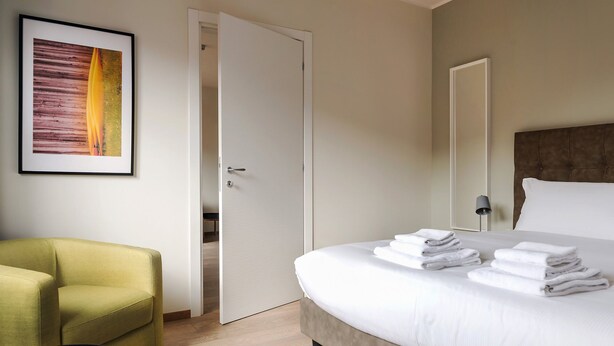 A hotel room with a bed, a green armchair, a door, and a framed picture on the wall. - Mi-brgz1c4 Bergonzoli Place - Bilo B - Canoe (Milan)