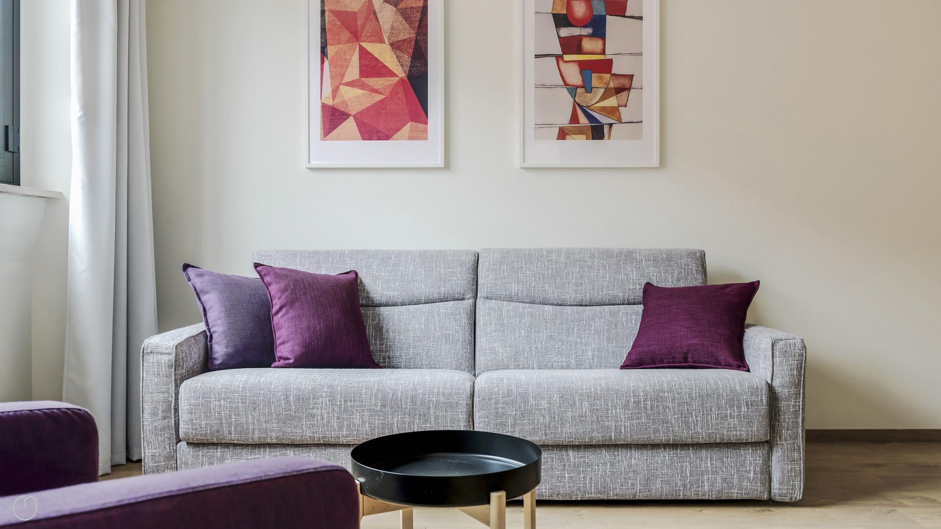 Ein graues Sofa mit violetten Kissen, einer schwarzen Ablage und abstrakten Bildern an der Wand.