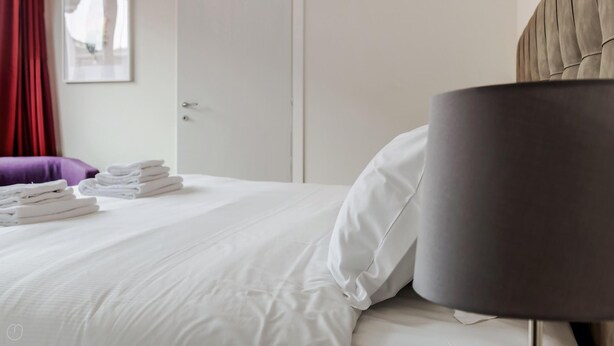 A neatly made bed with white linens, a stack of folded towels, a dark-colored headboard, and a closed door in a hotel room. - Mi-brgz1c3 Bergonzoli Place - Bilo B - Hugo (Milan)