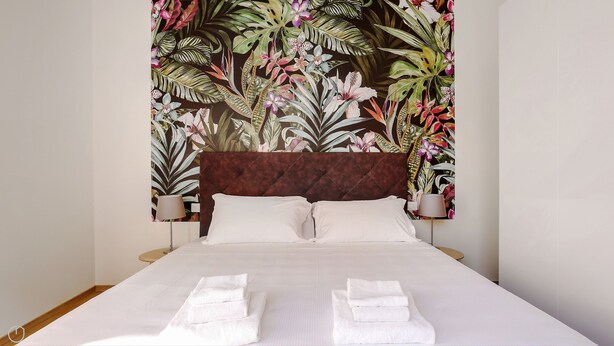 A hotel room with a bed, white linens, a dark headboard, and a tropical floral wall mural. - Mi-brgz1c1 Bergonzoli Place - Bilo B - Nina (Milan)