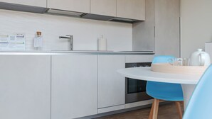 A modern kitchen with white cabinetry, a stainless steel range hood, and a built-in oven. There is a round table with a blue chair and a wooden cutting board. - Mi-brgz1b4 Bergonzoli Place - Terrace B - On the Grass (Milan)