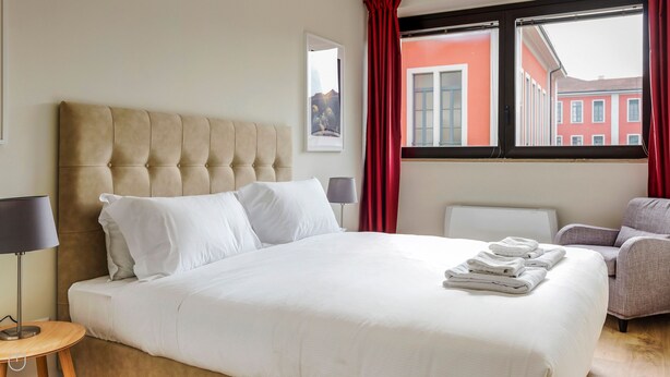 A hotel room with a large bed, a bedside table with a lamp, a chair, and a window with red curtains. - Mi-brgz1b3 Bergonzoli Place - Trilo - Shakespeare (Milan)