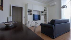 A modern living room with a dark wooden dining table, a black sofa, and a television mounted on a white cabinet. - Italianway Comfort - Lombardia 96 (Milan)