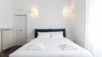 A neatly made bed with white linens and two folded towels, a white door, and a white radiator.