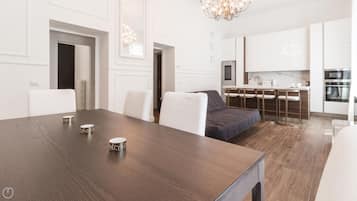 A modern kitchen with white cabinets, a dining area with a wooden table, and a bed with a dark blue bedspread.