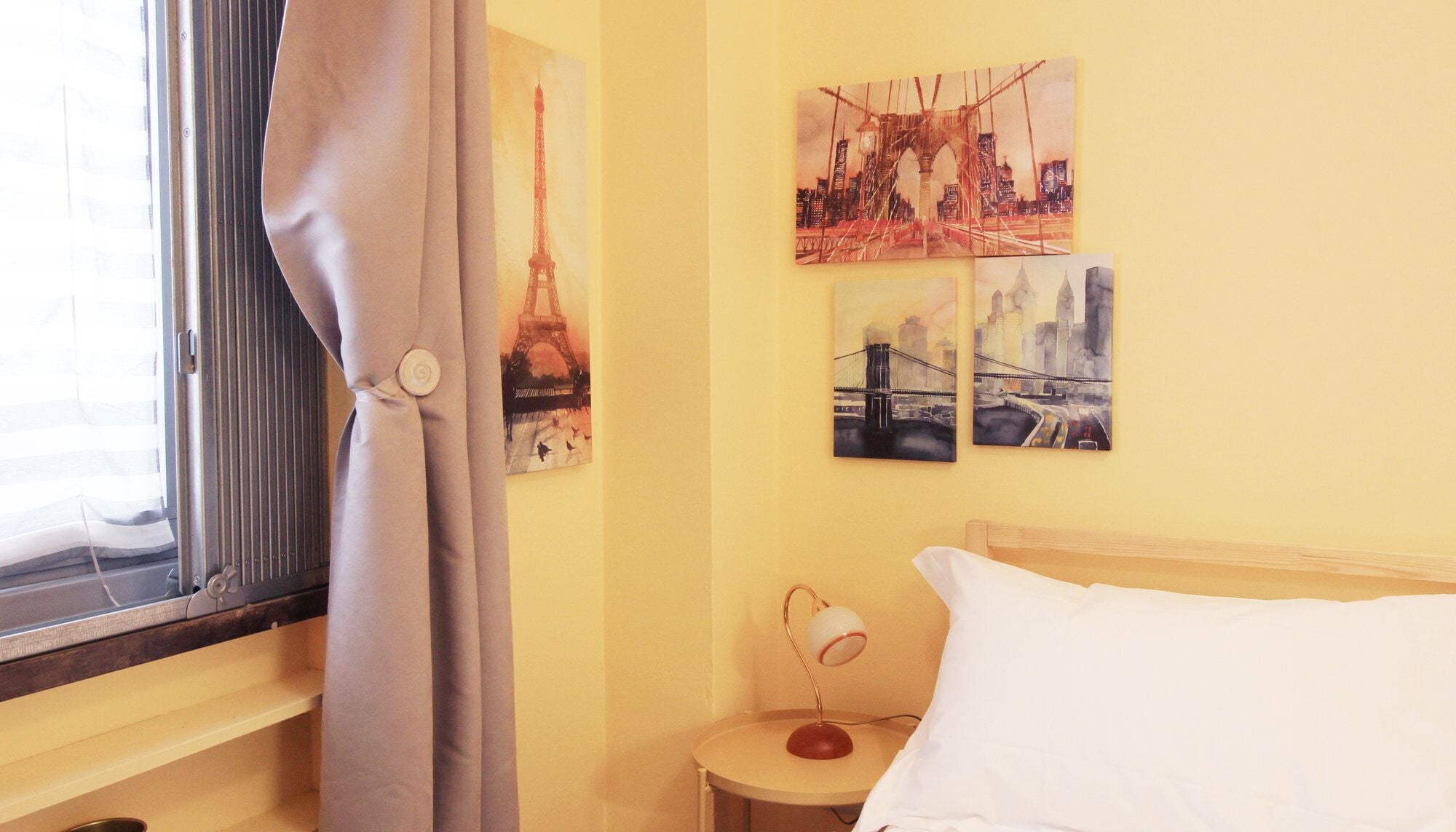 Habitación de hotel con cama, mesita de noche, lámpara y ventana con cortinas. Hay tres cuadros en la pared que representan puentes y paisajes urbanos.