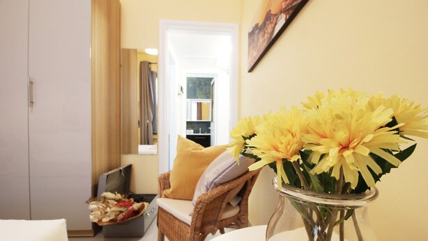 Een kamer met een rieten stoel, een tafel met een vaas vol gele bloemen en een witte kast.