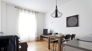 A modern, well-lit living room with a dining area, a TV, and a large window with curtains.