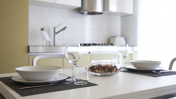 A modern kitchen with white cabinets, a stainless steel range hood, and a dining table set with a bowl, two wine glasses, and a glass of water. - Italianway Comfort - Spadolini 6 (Milan)