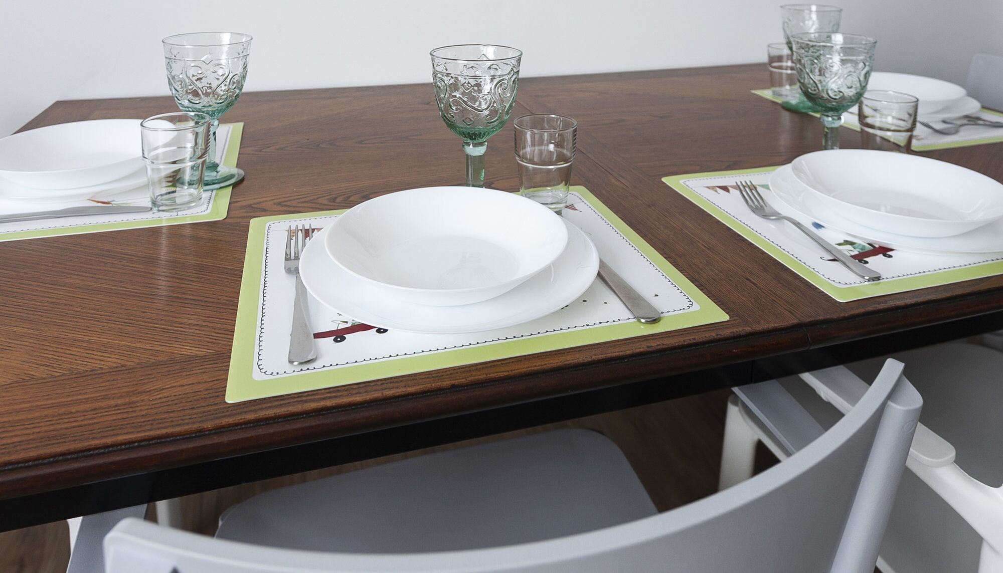 Ein Esstisch ist mit weißen Tellern, Besteck und grünem Glas auf einer Holzplatte mit grüner Tischdeko gedeckt.
