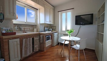 A compact kitchen with white cabinets, a brick backsplash, a small dining table with white chairs, and a TV mounted on the wall.