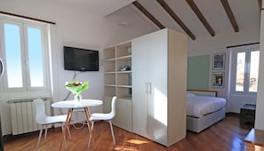 Una habitación con cama, televisión, armario blanco, una mesa con sillas y ventana.