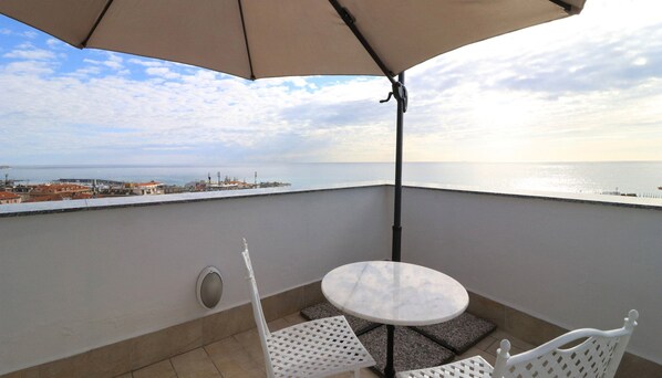 Un balcon avec une table et des chaises, un grand parasol et une vue sur la mer et les bâtiments.