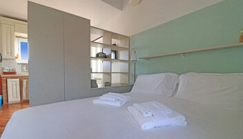 A bedroom with a bed, a built-in shelf unit, and a view of the kitchen.