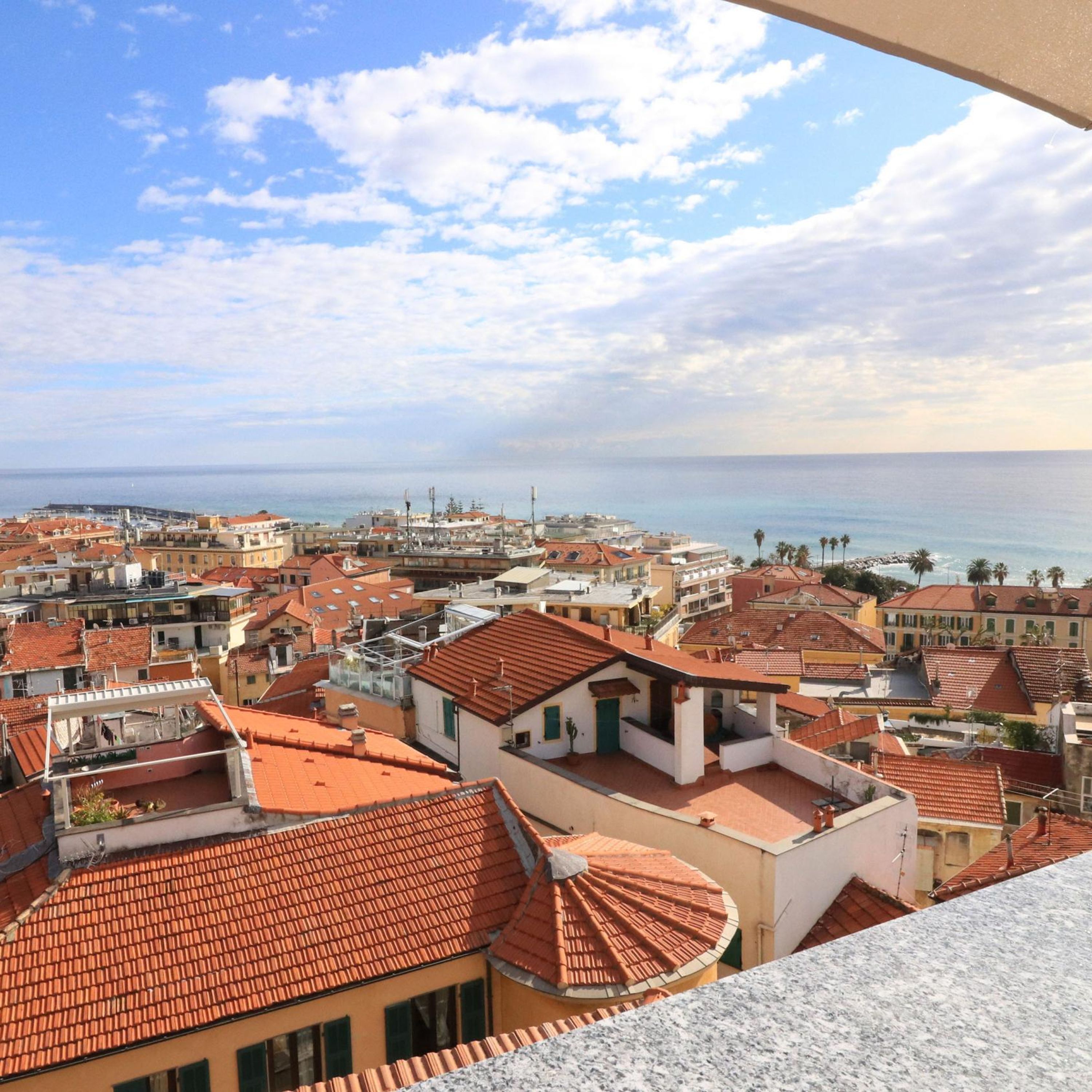 A coastal cityscape with red-tiled rooftops and buildings with balconies, overlooking the sea.