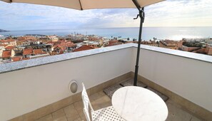 Un balcon avec une table et une chaise blanches, un parasol beige et une vue sur le paysage urbain de la côte.