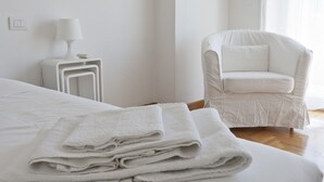 Un lit bien fait, avec des serviettes blanches pliées, une table de chevet blanche avec une lampe, et un fauteuil blanc dans une pièce minimaliste au revêtement de sol en bois.