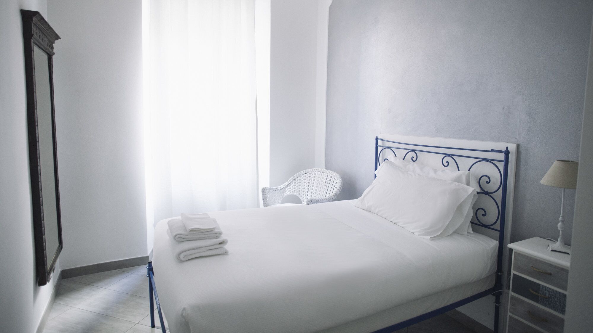 A neatly made bed with white linens, a white chair, and a bedside table with a lamp.