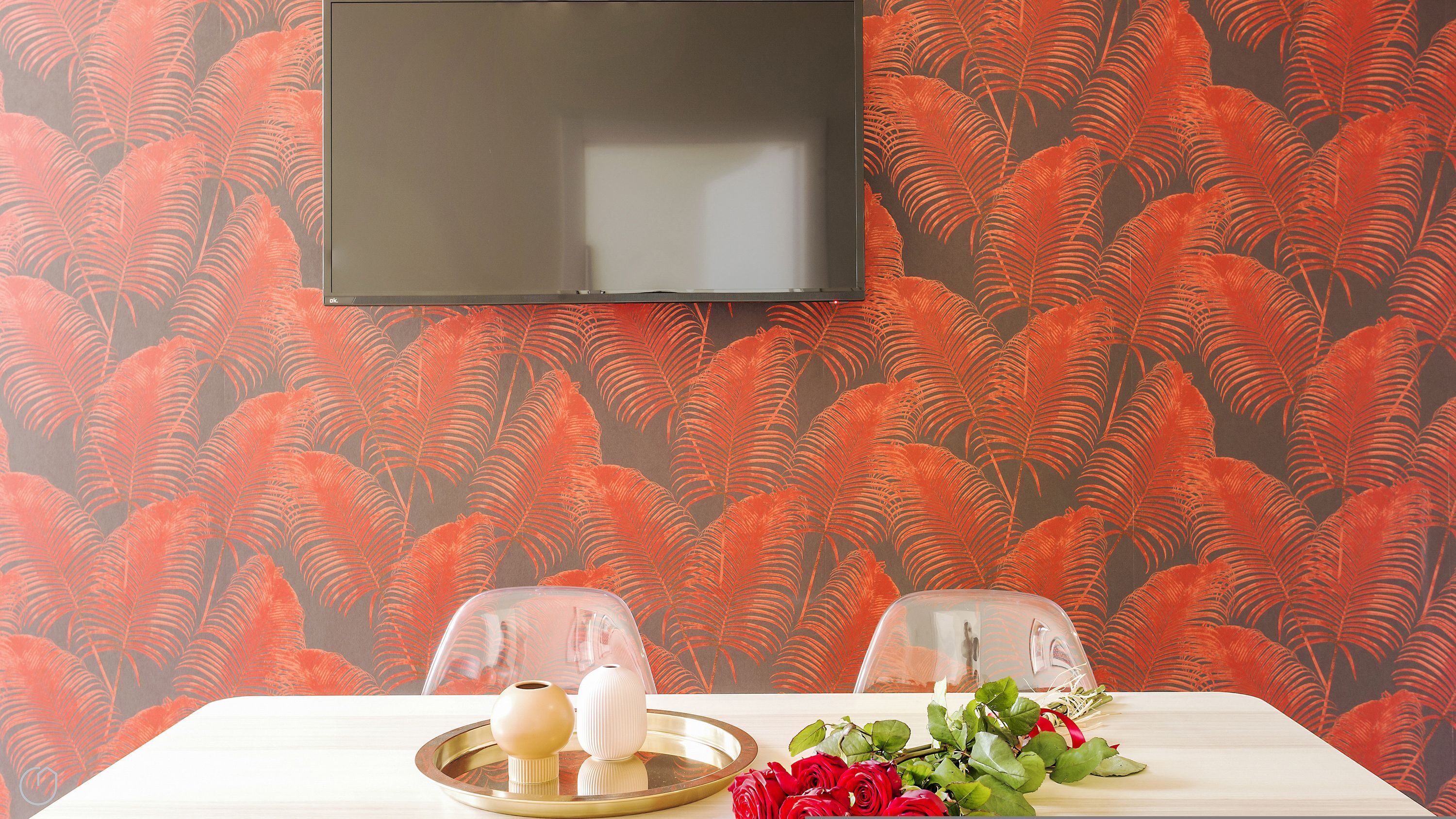 A room with a television mounted on the wall, a table with a vase and roses, and chairs with transparent covers.